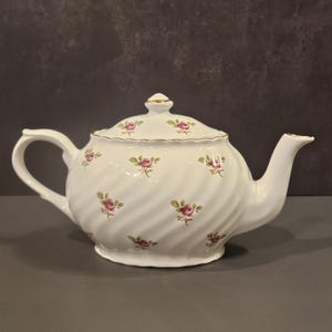 Puede incluir: Tetera de porcelana blanca con tapa, adornada con motivos de rosas rosas y hojas verdes. La tetera tiene un asa y un pico curvos, con ribetes dorados. El cuerpo presenta un diseño en espiral.