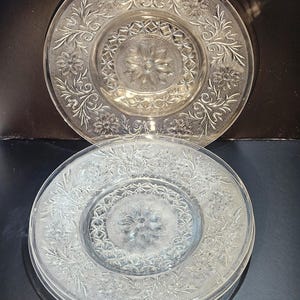 Puede incluir: Platos de vidrio transparente con diseños florales y de hojas en relieve. Los platos tienen un diseño floral central y un borde decorativo. Los platos están apilados, con un plato encima y varios debajo.