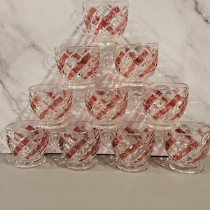 May include: A collection of ten clear glass teacups with a red diamond pattern. Each cup has a handle and sits on a small footed base. The cups are stacked in a pyramid shape, showcasing their design.