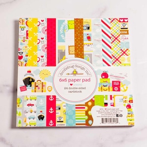 May include: A 6x6 inch paper pad with a travel theme, featuring 24 double-sided cardstock sheets. The cover displays colorful patterns with travel-related illustrations like suitcases, airplanes, and stamps. The pad is from Doodlebug Design Inc.
