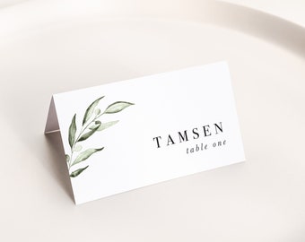 Op de afbeelding: Een witte naambordje met de naam "Tamsen" en "table one" in het zwart gedrukt. De kaart heeft een aquarel illustratie van groene olijftakken. De kaart is gevouwen en op een wit bord geplaatst.