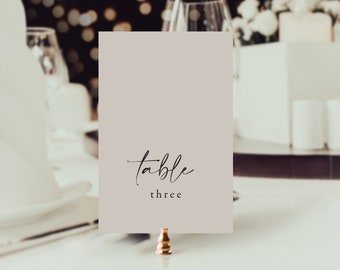 May include: A rectangular, light taupe table number card with the word "table" in elegant script and "three" below. The card is held by a small, gold-colored metal clip. The background is blurred, suggesting a wedding or event setting.
