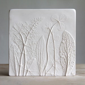 May include: White ceramic wall art featuring a raised design of pressed leaves and flowers. The design is detailed and intricate, showcasing the veins and textures of the plants.