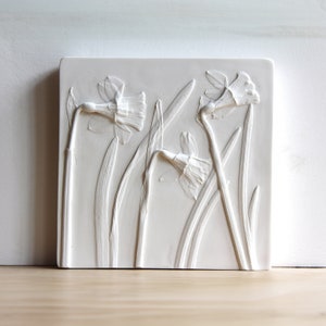 May include: A square, off-white relief sculpture featuring three daffodils with long, slender leaves. The flowers are in various stages of bloom, with detailed petals and stems. The artwork is mounted on a square base, creating a three-dimensional effect.