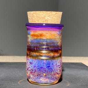 May include: A small glass jar with a cork stopper featuring an iridescent finish. The body of the jar displays a gradient of colors, transitioning from a deep purple rim to horizontal bands of orange and gold, ending in a speckled blue base.