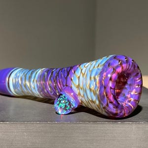May include: A glass pipe featuring a swirling pattern of purple and light blue colors with a metallic gold finish. The cylindrical body has a flared opening at one end and a small, circular, iridescent blue accent piece attached to the side.