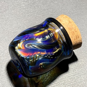 May include: A decorative glass jar with a cork stopper. The jar features a swirling pattern of blue, gold, and red colors. The jar is designed with a rounded shape and a black rim.