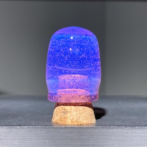 Op de afbeelding: Een gloeiende blauw-paarse flesstopper met een kurken basis. De stopper heeft een afgeronde bovenkant en een cilindrisch lichaam. Het blauwe gedeelte lijkt doorschijnend, met kleine, lichtgekleurde insluitsels. De basis is gemaakt van natuurlijk kurk.