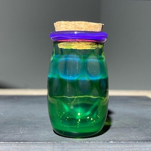 Op de afbeelding: Een klein glazen flesje met een levendig groen lichaam met een kuiltjesachtig, getextureerd oppervlak. De hals van het flesje is geaccentueerd met een felpaarse glazen ring en de opening is afgesloten met een lichtbruine, cilindrische kurk.