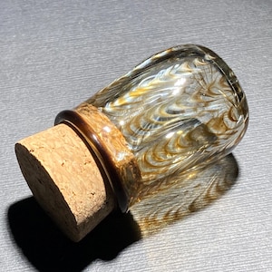 May include: A decorative glass bottle with a cork stopper. The bottle has a clear body with swirling gold and brown patterns. The cork is a light brown color, and the bottle has a dark brown rim.