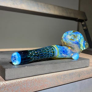 May include: A hand-blown glass pipe with a blue, green, and yellow color scheme. The bowl and mouthpiece are rounded, while the stem has a textured, honeycomb-like pattern. The pipe is resting on a dark gray surface.