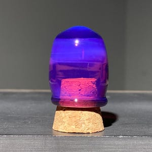 Op de afbeelding: Een klein, doorschijnend paars glazen koepelvormig object dat rust op een lichtbruine, getextureerde kurken basis. Het paarse glas heeft een gladde, afgeronde bovenkant en een helder, cilindrisch onderste gedeelte op de ronde kurken stop.