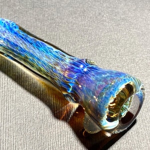 Op de afbeelding: Een glazen pijp met een blauwe en gouden iriserende afwerking. De pijp is ongeveer 10 cm lang en heeft een afgeronde kom.