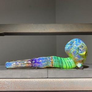 May include: A glass smoking pipe featuring a swirling pattern of vibrant blue, green, and iridescent colors. The bowl end is rounded with a marbled design, while the stem transitions into a deep blue hue with a shimmering, reflective finish.