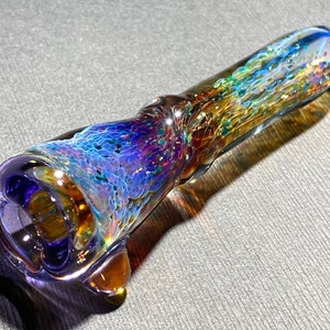 Può includere: Una colorata pipa di vetro con una coppa svasata e un corpo lungo e cilindrico. La pipa presenta una miscela di blu, viola e oro, con un motivo strutturato e maculato. Il vetro è traslucido ed è lungo circa 15 cm.