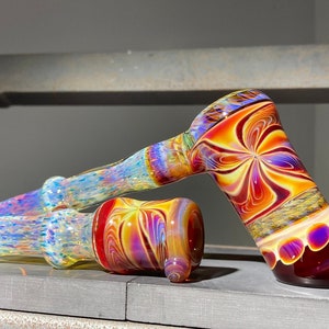 May include: A colorful glass pipe with a red, yellow, and purple swirl design. The pipe has a round bowl and a long, thin stem.