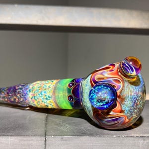 May include: A decorative glass pipe with a vibrant, multi-coloured design. The pipe features a bulbous bowl with a large blue orb and a smaller orange orb. The stem is long and cylindrical, with a mix of blue, purple, yellow, green, and orange.