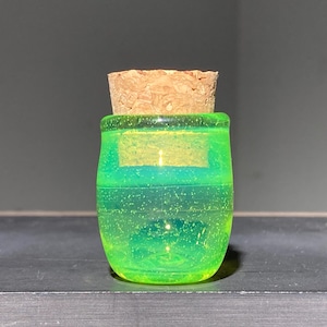 Op de afbeelding: Een kleine, ronde, heldere glazen pot met een kurken stopper. De pot is gevuld met een heldere, neon groene vloeistof. De pot staat op een donkere ondergrond tegen een grijze achtergrond. De kurk is lichtbruin.