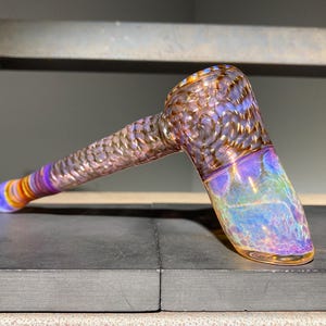 May include: A hand-blown glass pipe featuring a textured, iridescent finish with swirling patterns in shades of purple, blue, and gold. The stem has a tapered design leading to a flared mouthpiece, while the bowl displays a reflective, metallic sheen.