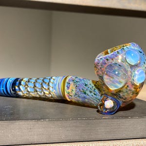 May include: A colorful glass pipe with a blue, yellow, and purple design. The pipe features a bowl with three large, round, opalescent accents. The stem has a scale-like pattern in blue and gold. The overall design is artistic and decorative.
