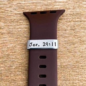 May include: A dark brown Apple Watch band with a silver metal accent. The metal accent has the text "Jer. 29:11" engraved on it. The band is made of a flexible material and has multiple holes for adjustment.