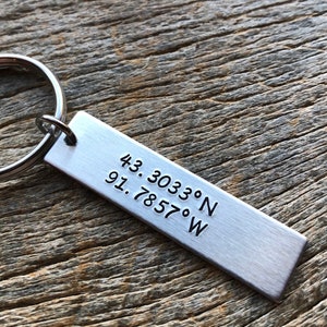 May include: Silver keychain with a rectangular tag engraved with the coordinates "43.3033°N 91.7857°W".