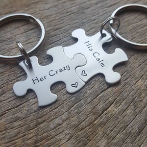 May include: Two silver-colored metal puzzle piece keychains with circular rings. One piece is stamped with the text Her Crazy and a small heart, while the other piece is stamped with the text His Calm and a small heart. The pieces interlock together.
