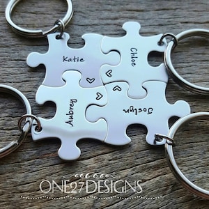 May include: Four silver puzzle piece keychains with the names Katie, Chloe, Aubrey, and Jocelyn engraved on them. Each piece has a small heart engraved on it.