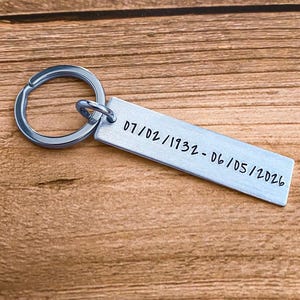 May include: A rectangular silver-toned metal keychain tag featuring a brushed finish and a circular split ring. The tag is engraved with the black text 07/02/1932 - 06/05/2026, indicating a date range in a simple, sans-serif font style.