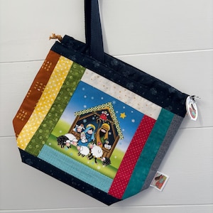 May include: A handmade tote bag with a Nativity scene. The bag features a patchwork design with various fabric patterns and colors, including brown, yellow, green, and blue. The main image depicts Mary, Joseph, and baby Jesus in a stable with sheep.