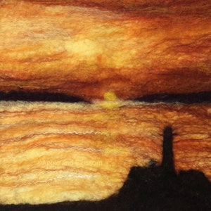 May include: A needle felted artwork depicting a sunset over a body of water with a silhouette of a lighthouse in the foreground. The sky is a mix of orange, yellow, and red, with white clouds. The water is a deep blue, with white waves. The lighthouse is a tall, slender structure with a white light at the top.