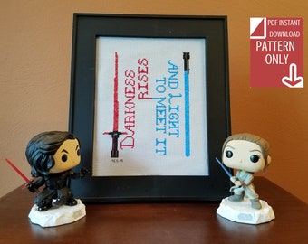 May include: A framed cross-stitch pattern with the text "Darkness Rises To Meet It And Light" and two lightsaber images, one red and one blue. The frame is on a wooden table with two Star Wars character figurines, one with a red lightsaber and one with a blue lightsaber.