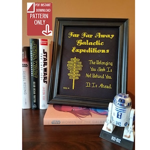 May include: A framed cross-stitch design with the text "Far Far Away Galactic Expeditions" and a quote from Star Wars: "The Belonging You Seek Is Not Behind You. It Is Ahead." The design is in gold thread on a black background. A Star Wars R2-D2 figurine is in the foreground.