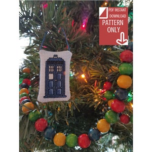 May include: A white fabric ornament with a blue and white cross-stitch design of a TARDIS, a time-traveling police box from the Doctor Who television series. The ornament is hanging on a Christmas tree with a string of colorful wooden beads.