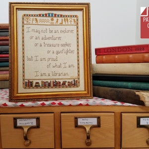 Puede incluir: Un bordado enmarcado con el texto "Puede que no sea un explorador o un aventurero o un buscador de tesoros o un pistolero, pero estoy orgulloso de lo que soy. Soy bibliotecario." El marco es dorado y el bordado está sobre tela de color crema. Hay libros apilados sobre una mesa de madera con cajones etiquetados como "Nefertiti Ptolomeo VI", "54T4- Tuth Mos" y "Jerjes- Z".