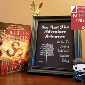 May include: A framed cross-stitch pattern with the text "Ice And Fire Adventure Getaways" and a signpost with locations from the book series "A Song of Ice and Fire". The text "Winter Is Coming. Book Your Vacation Today." is also included. A book titled "Fire & Blood" by George R.R. Martin is visible in the background.