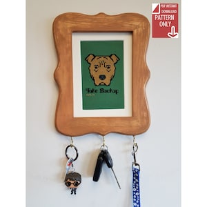 May include: A wooden key holder with a framed cross-stitch of a dog's face and the text "Take Backup". The frame has a decorative, ornate design. Three key rings hang from the holder, one with a small figurine, one with car keys, and one with a blue leash.