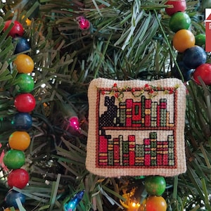 May include: A cross-stitch ornament featuring a bookshelf filled with colorful books. A black cat sits on the top shelf. The ornament is hanging on a Christmas tree with a string of colorful beads.