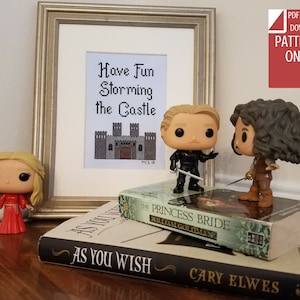 May include: Cross stitch pattern featuring a castle with the text "Have Fun Storming the Castle". The pattern is displayed in a framed picture with a white background. The image also includes three small figurines of characters from the movie "The Princess Bride" and two books with the titles "The Princess Bride" and "As You Wish".