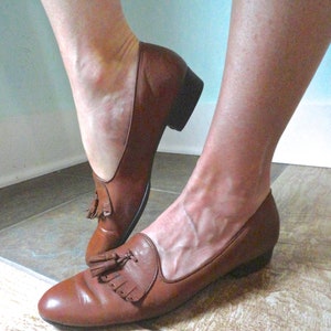 May include: Brown leather loafers with a low heel and tassel detail. The shoes have a classic design and a polished finish. The loafers are a slip-on style and appear to be well-worn.