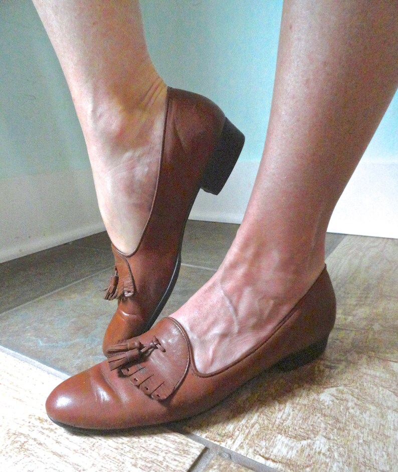 May include: Brown leather loafers with a low heel and tassel detail. The shoes have a classic design and a polished finish. The loafers are a slip-on style and appear to be well-worn.