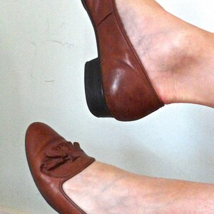 May include: A pair of brown leather flats with a small heel. The shoes feature a tassel detail and a rounded toe. The shoes are a warm brown colour and appear to be well-worn.