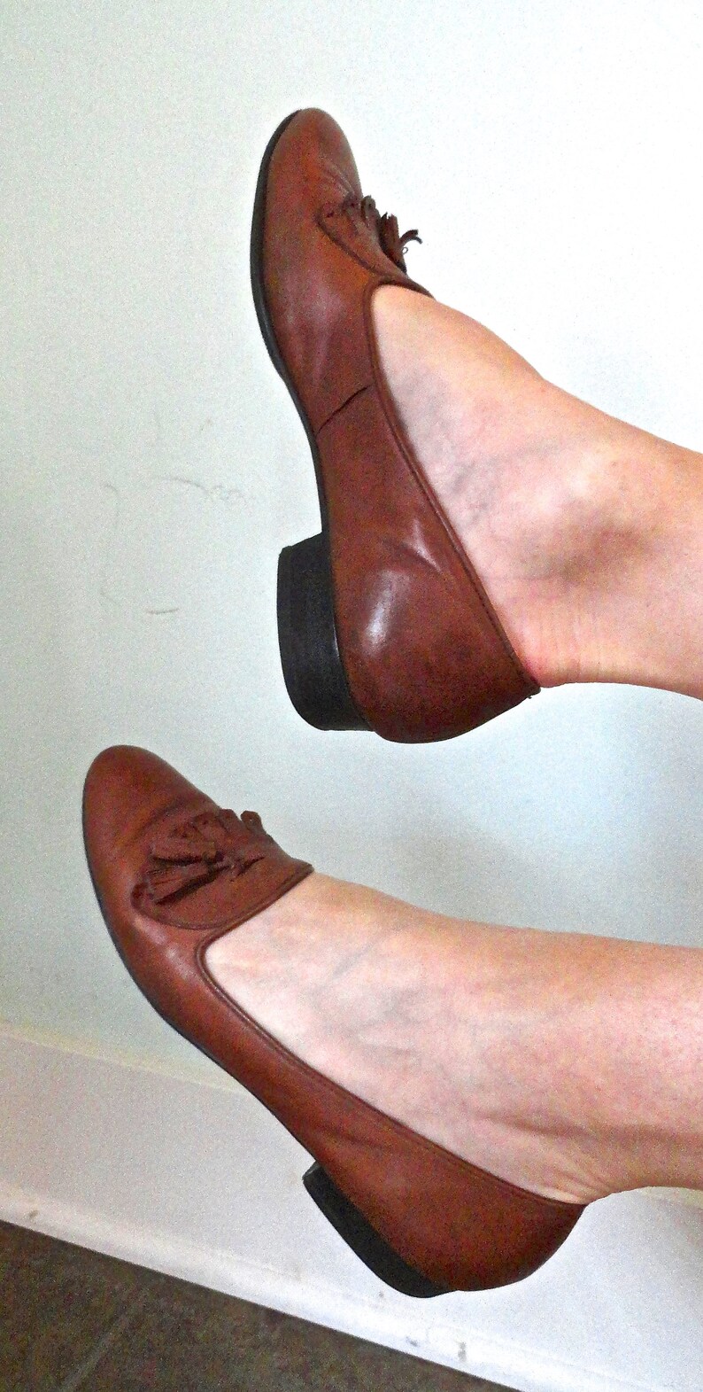 May include: A pair of brown leather flats with a small heel. The shoes feature a tassel detail and a rounded toe. The shoes are a warm brown colour and appear to be well-worn.