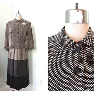 May include: A vintage two-piece suit displayed on a mannequin. The jacket is speckled gray with a rounded collar and button closure. The skirt is pleated, transitioning from gray to black. A white flower brooch with a pearl center adorns the jacket.