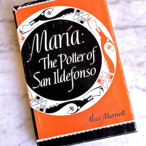May include: A vintage book titled "Maria: The Potter of San Ildefonso" by Alice Marriott. The book has an orange cover with a black and white circular design and text. The book is set against a marble background.