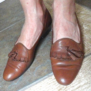 May include: Brown leather loafers with tassel detailing. The shoes have a rounded toe and a low heel. The leather appears slightly worn, giving a vintage look. A classic style of shoe, suitable for many occasions.