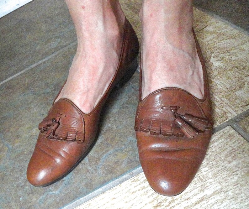 May include: Brown leather loafers with tassel detailing. The shoes have a rounded toe and a low heel. The leather appears slightly worn, giving a vintage look. A classic style of shoe, suitable for many occasions.
