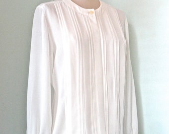 May include: A white blouse with long sleeves and a mandarin collar. The front of the blouse features vertical pleats and a single button closure. The sleeves have buttoned cuffs. The blouse is displayed on a mannequin.