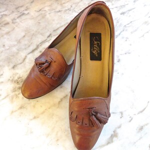 May include: A pair of brown leather loafers with tassel detailing. The shoes have a rounded toe and a low heel. The brand name "Selly" is visible on the insole. The shoes are displayed on a marble surface.
