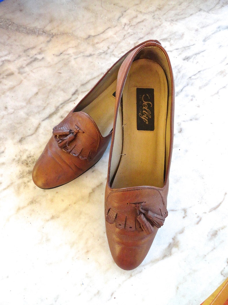 May include: A pair of brown leather loafers with tassel detailing. The shoes have a rounded toe and a low heel. The brand name "Selly" is visible on the insole. The shoes are displayed on a marble surface.